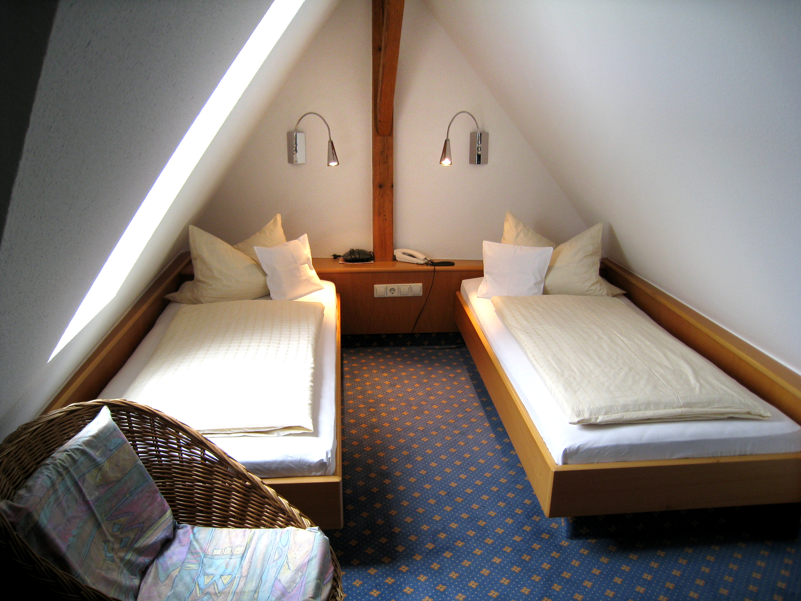 Comfort twin bedroom with two single beds separated by a wooden nightstand under a slanted ceiling and a wicker chair in the corner